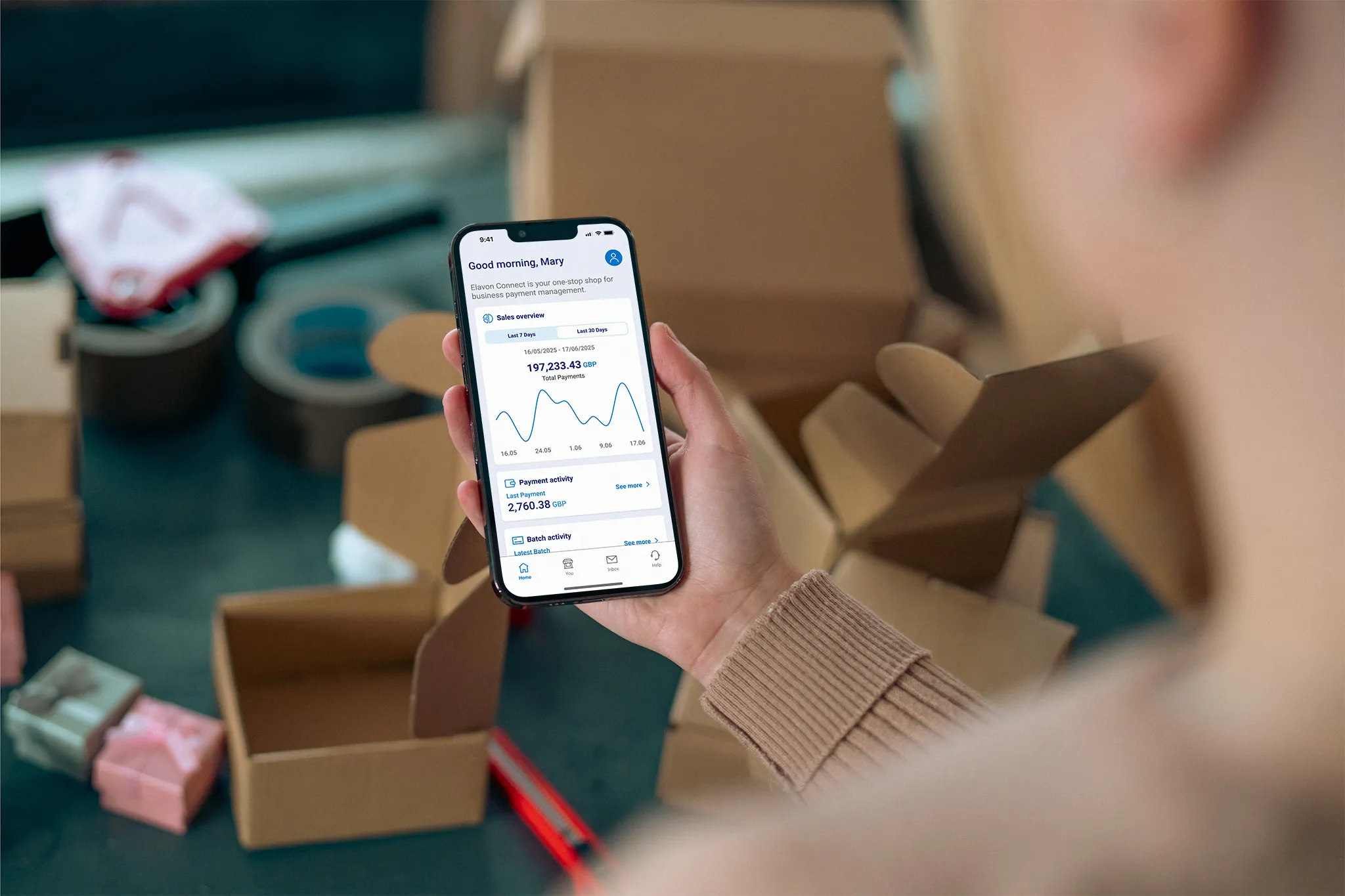 A person holding a smartphone displaying Elavon Connect with the greeting 'Good morning, Mary!' visible on the screen. In the background, there are cardboard boxes, tape, and small wrapped items, suggesting a packing or shipping environment.
