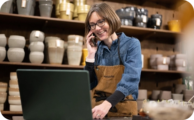 Shop keeper in store holding phone to ear behind a counter with a laptop on counter